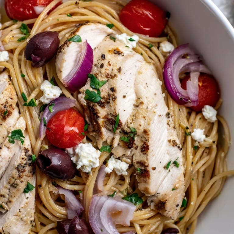 A close-up of Greek Chicken Spaghetti reveals juicy tomatoes, briny olives, and creamy feta cheese in a vibrant serving.
